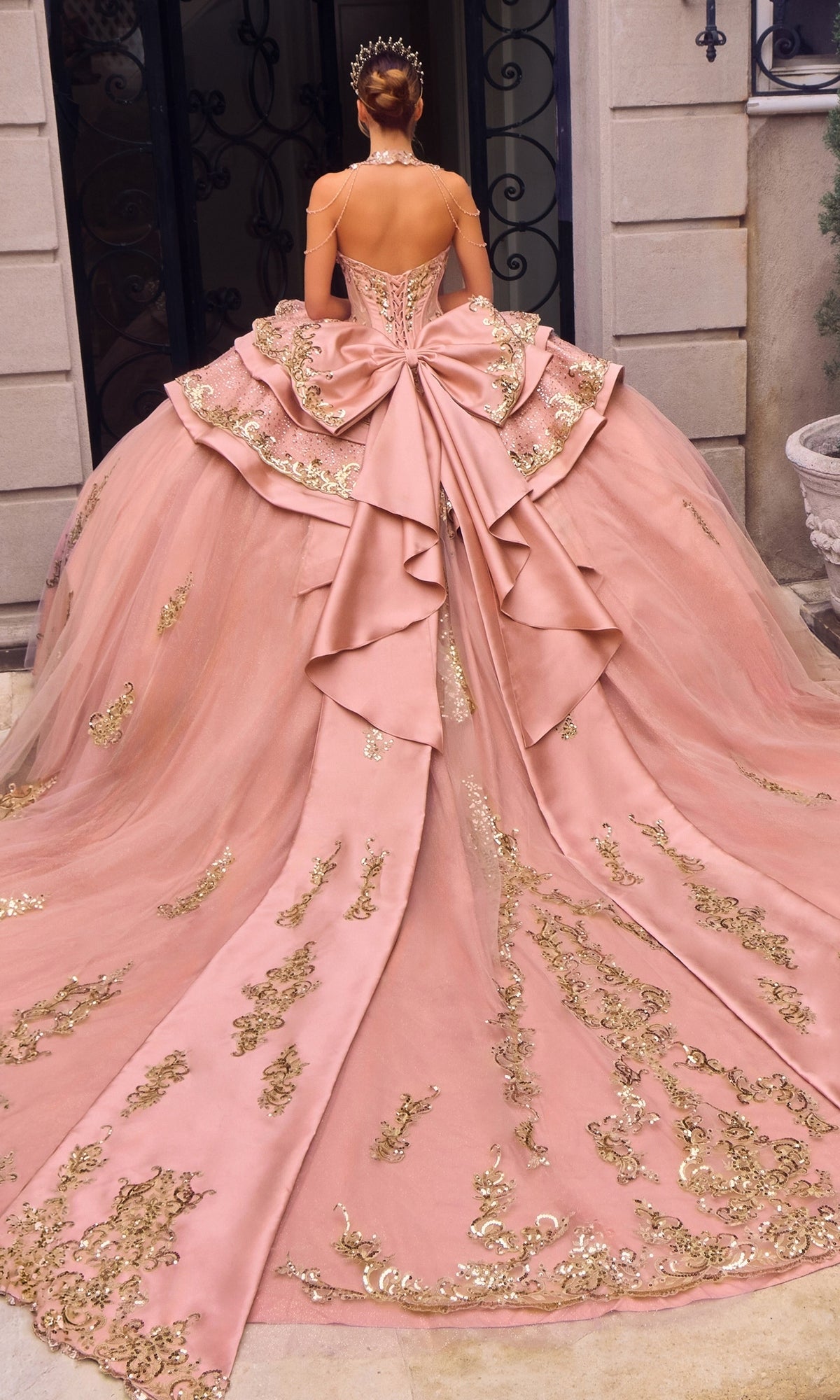 A woman in the Quinceanera Dress 15744 By Ladivine, featuring a pink ballgown silhouette with gold embroidery and a large bow, stands facing away in front of an ornate building entrance.